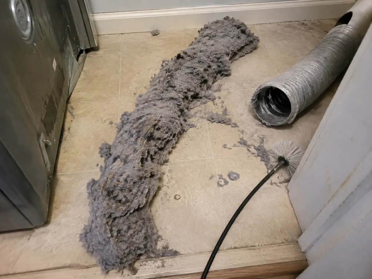A long rope of packed lint on a laundry room floor beside a cleaning brush
