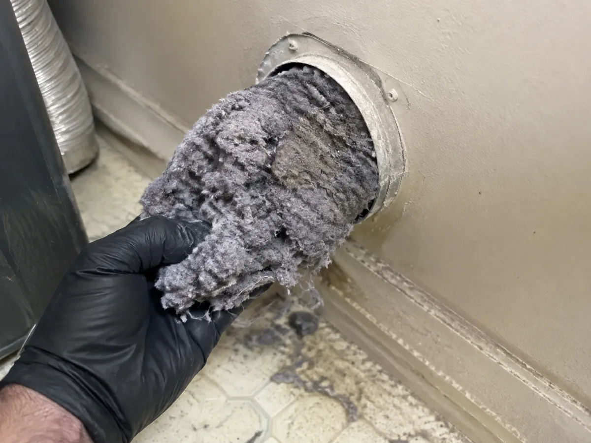 A gloved hand pulling a thick plug of lint out of a dryer vent in the wall